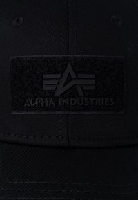 Crna kapa s teksturiranim aplikacijom koja sadrži logo "ALPHA INDUSTRIES" u podebljanim, utisnutim slovima. Glatka tkanina i strukturirani oblik.