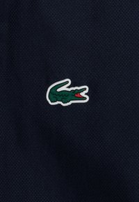 Tamsiai mėlynas tekstūruotas audinys su ryškiai žaliu ir raudonu krokodilo logotipo pleistru, nubrėžtu baltai, esantis kairėje pusėje.