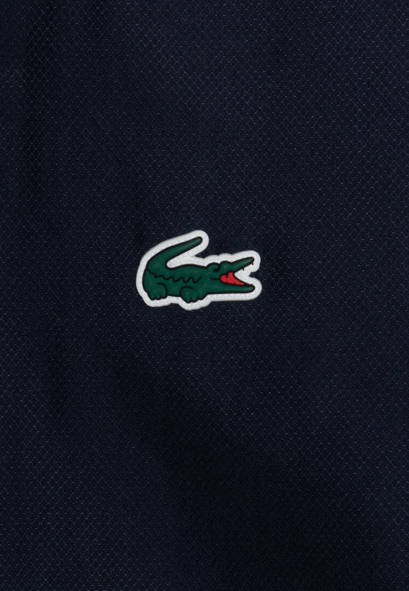 Marineblauer strukturierten Stoff mit prominentem grünen und roten Krokodil-Logo-Patch, umrandet in Weiß, links positioniert.