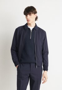Mlad moški s kratkimi kodrastimi lasmi, oblečen v mornarsko modro jakno, mornarsko modro zip-up pulover in usklajene mornarsko modre hlače, stoji pred belo ozadjem.