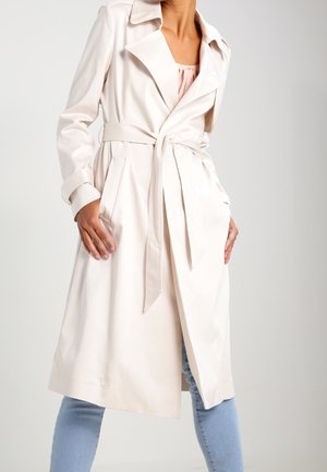 Kvinde iført en lys beige trenchcoat med bælte over en sart lyserød top og lyseblå jeans, stående foran en ensfarvet baggrund.