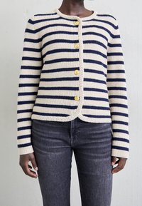 Raidallinen villatakki, jossa on kerma ja laivastonsininen, kultaiset napit, ribattu pinta, pyöreä pääntie ja lyhennetty helma. Yhdistetty tummiin farkkuihin.