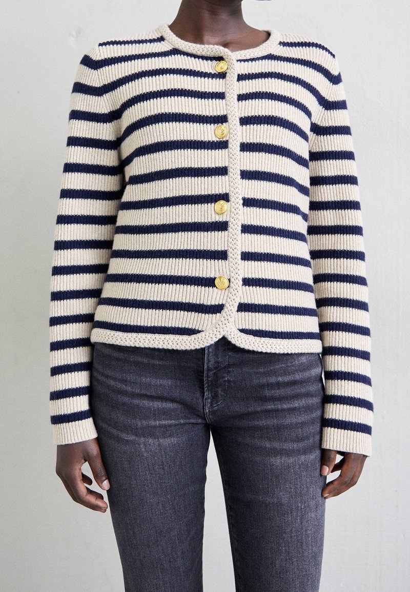 Raidallinen villatakki, jossa on kerma ja laivastonsininen, kultaiset napit, ribattu pinta, pyöreä pääntie ja lyhennetty helma. Yhdistetty tummiin farkkuihin.