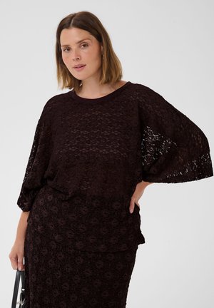 Femme aux cheveux mi-longs portant un haut en dentelle foncé à larges manches et une jupe assortie, tenant un sac à main noir.