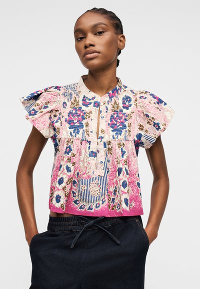 Femme portant un chemisier à manches courtes à motifs floraux avec des épaules froncées et un pantalon sombre à cordon, debout les mains dans les poches.