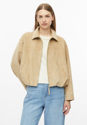 Vrouw met steil lichtbruin haar draagt een beige suède jas met rits over een licht shirt en blauwe spijkerbroek tegen een witte achtergrond.