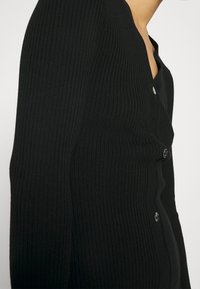 Cardigan noir côtelé avec col en V, orné de boutons décoratifs à l'avant et d'une coupe ajustée qui met en valeur la silhouette.