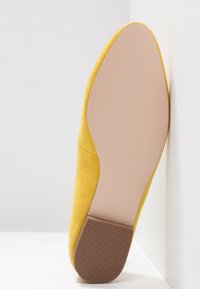 Chaussure plate en daim jaune avec un bout arrondi et une semelle en caoutchouc marron texturée, appuyée contre un fond neutre.