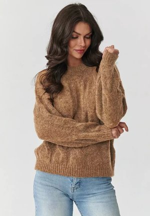 Brązowy sweter dzianinowy z teksturowanym wzorem, w oversize'owym fasonie i ściągaczami przy mankietach. Noszony z jasnoniebieskimi dżinsami. Miękki, puszysty materiał.