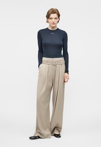 Γυναίκα που στέκεται φορώντας εφαρμοστή σκούρα μπλούζα με μακριά μανίκια και φαρδιά μπεζ παντελόνια ψηλόμεσα με ζώνη που ταιριάζει και μαύρα παπούτσια.