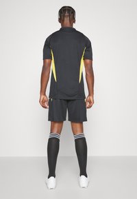 Camiseta y pantalones cortos atléticos negros con detalles amarillos. Tres rayas en los calcetines negros. Textura suave, mangas cortas, diseño ajustado.