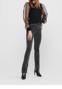 Haut noir à manches bouffantes en voile transparent, associé à un jean slim gris foncé et des escarpins noirs texturés, mettant en avant un design moderne et une silhouette élégante.