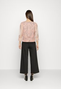 Blusa rosa blush com mangas bufantes transparentes, detalhada com lantejoulas florais; combinada com calças largas de cintura alta pretas e sapatos de salto chunky.