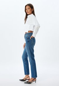 Valkoinen lyhyt nappisaumapaita etutaskulla, yhdistettynä sinisiin suoriin farkkuihin ja mustiin kärki-slippereihin.