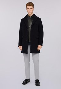 Cappotto di lana nera con colletto a scialle, design a un petto, tasche profonde, indossato sopra una giacca di pelle scura, abbinato a pantaloni grigio chiaro e scarpe nere.