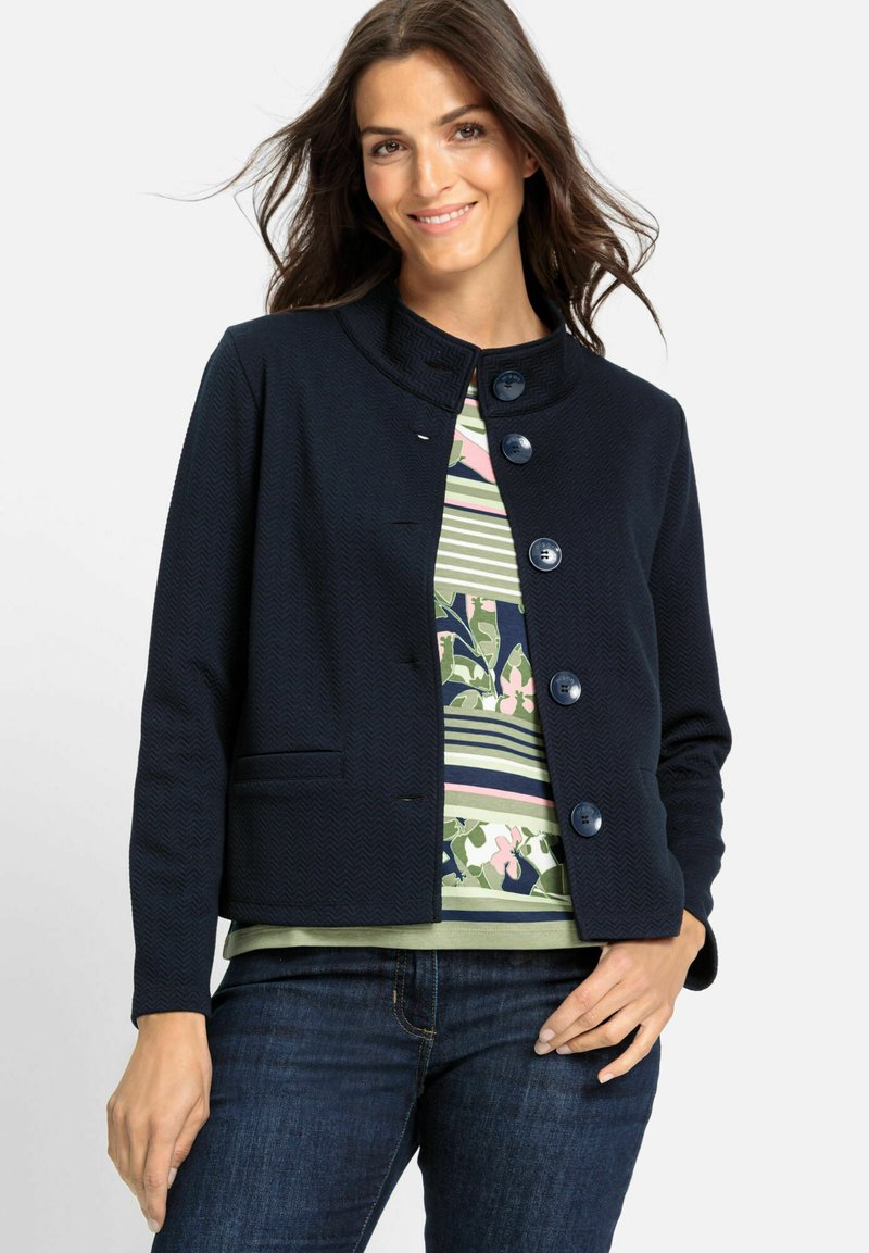 Marineblaue Jacke mit einem strukturierten Fischgrätenmuster, runden Knöpfen und einem runden Kragen, kombiniert mit einem gestreiften Blumenshirt.