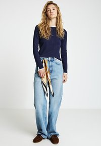 Marineblaue Langarmbluse mit weißen Kragenakzenten, hellblaue weite Jeans und ein gemustertes Halstuch, das um die Taille gebunden ist, getragen mit braunen Schuhen.