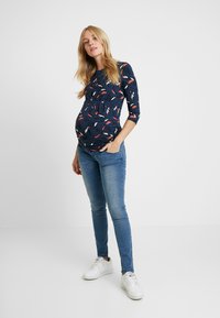Top de maternidade em azul marinho com estampa de penas, com mangas de três quartos. Combinado com jeans skinny azuis e ténis brancos.