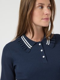Femme portant un pull côtelé bleu marine avec un col à volants présentant des rayures blanches et trois boutons blancs sur le devant.