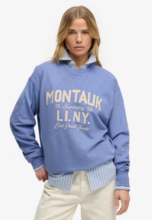 Vrouw met blond haar, gekleed in een blauwe Montauk-trui over een gestreept overhemd en lichtgekleurde broek, poseert met een hand op haar heup.