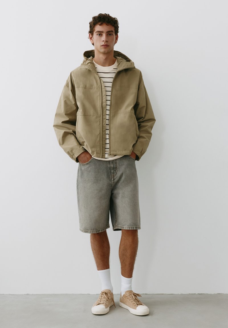 Jeune homme debout portant une veste à capuche beige, un t-shirt rayé, un short en jean gris, des chaussettes blanches et des baskets beiges devant un mur blanc.