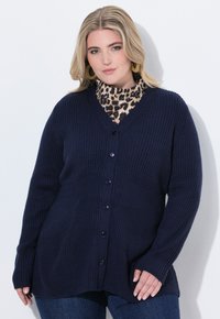 Mörkblå ribbad cardigan med V-hals, försedd med sex knäppningar, långa ärmar och en avslappnad passform. Bärs över en turtleneck med leopardmönster.