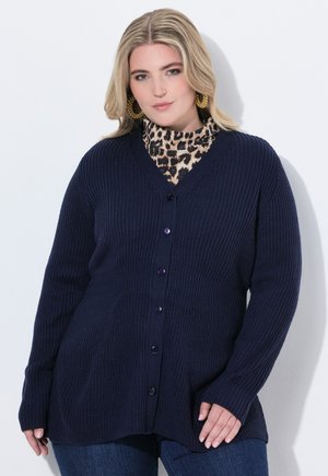 Cardigan côtelé bleu marine avec un décolleté en V, doté de six boutons, de manches longues et d'une coupe décontractée. Porté par-dessus un col roulé à imprimé léopard.