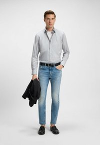 Chemise à rayures gris clair, portée avec un jean bleu clair. Ceinture noire et mocassins. Veste noire tenue dans la main gauche.