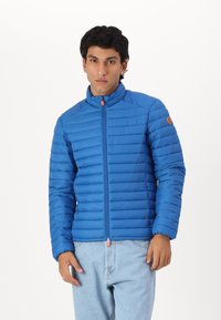 Blauwe pufferjack met horizontaal gestikte ontwerpen, hoge kraag, voorrits met oranje trekkoord en een logo-opdruk op de linker mouw.