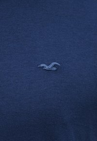 Μικρό κεντημένο λογότυπο γλάρου στο κέντρο ενός πλεκτού υφάσματος σε σκούρο μπλε, πιθανώς ενός μπλουζιού ή φούτερ.