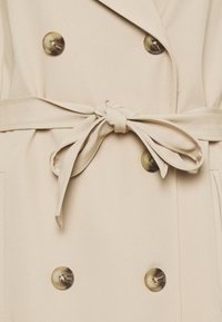 Beige double-breasted trench coat featuring large buttons, a knotted fabric belt, and side pockets. Smooth texture with a tailored fit.