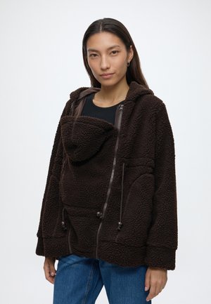 Frau trägt dunkelbraune Fleecejacke mit Fronttasche und Kapuze, kombiniert mit blauer Jeans, steht vor weißem Hintergrund.
