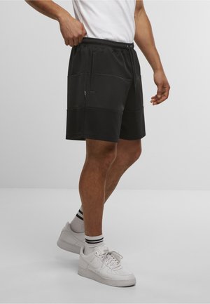 Mann iført svarte knelengde shorts, hvite crew-sokker med svarte striper og hvite Nike-sneakers, stående foran en ensfarget lys bakgrunn.