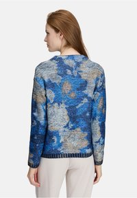 Frau, die mit dem Rücken zur Kamera steht, trägt einen blau-beigen, floral gemusterten Strickpullover und hellbeige Hose vor einem schlichten Hintergrund.