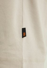 Beiger Baumwollstoff mit gerippter Textur, versehen mit einem schwarzen und braunen Etikett an der Seitennaht und sichtbaren Nähdetails.