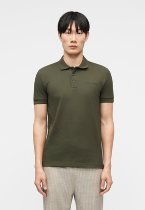 Νέος άνδρας που φοράει σκούρο πράσινο πόλο με κοντά μανίκια και ανοιχτό γκρι παντελόνι με καρό σχέδιο, στέκεται μπροστά σε ένα απλό λευκό φόντο.