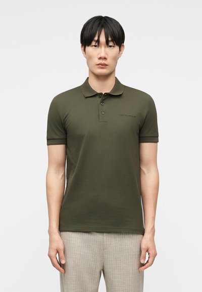 Jeune homme portant un polo à manches courtes vert foncé et un pantalon gris clair à carreaux, debout devant un fond blanc uni.