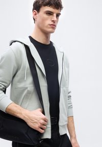 Νέος άνδρας που φοράει ανοιχτό γκρι φούτερ με φερμουάρ και μαύρο μπλουζάκι, κρατώντας μία μαύρη τσάντα χιαστί με ιμάντα στον ώμο του.