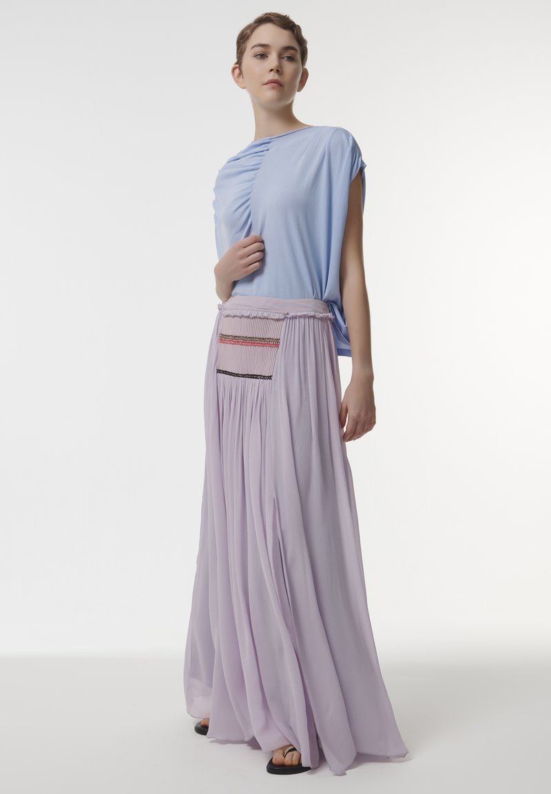 Lichtblauw drapeertop met korte mouwen, gecombineerd met een lila plissé-maxirok met een smocktail en decoratieve stiksels.