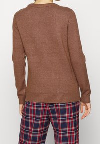 Pull en maille marron avec un col rond, des manches longues et un ourlet côtelé. Associé à un pantalon à carreaux dans des tons rouges, bleu marine et jaunes.