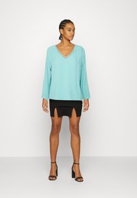 Blusa turquesa de mangas longas com decote em V e um acabamento decorativo, combinada com uma saia mini preta com uma fenda frontal, usada com saltos pretos.