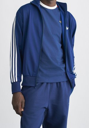 Mężczyzna noszący granatowy dres Adidas z białymi paskami na rękawach oraz pasującą granatową koszulkę pod spodem.
