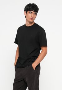 Black cotton t-shirt with a round neck, short sleeves, and a small logo detail on the chest, worn with dark pants.
