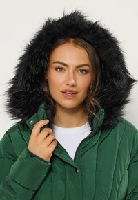 Grön puffjacka med en svart fuskpälsfodrad huva, med dragkedja framtill och synlig quiltad struktur. Modellen håller i jackans krage.