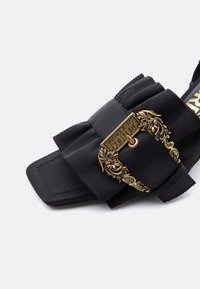 Μαύρο σανδάλι Versace Jeans Couture με ραμμένη λουρίδα και περίτεχνη χρυσή αγκράφα με λεπτομέρειες λουλουδιών και χαραγμένο λογότυπο.