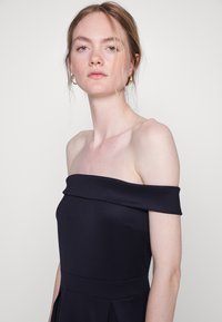 Vestido azul marinho com ombros à mostra, com um corpete justo, saia plissada e textura suave. Apresenta um decote largo e drapeado e elementos de design mínimos.