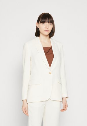 Weißer taillierter Blazer mit einem einzelnen goldenen Knopf, zwei Fronttaschen und glattem Stoff. Über einem braunen Oberteil mit einfachem Ausschnitt getragen.