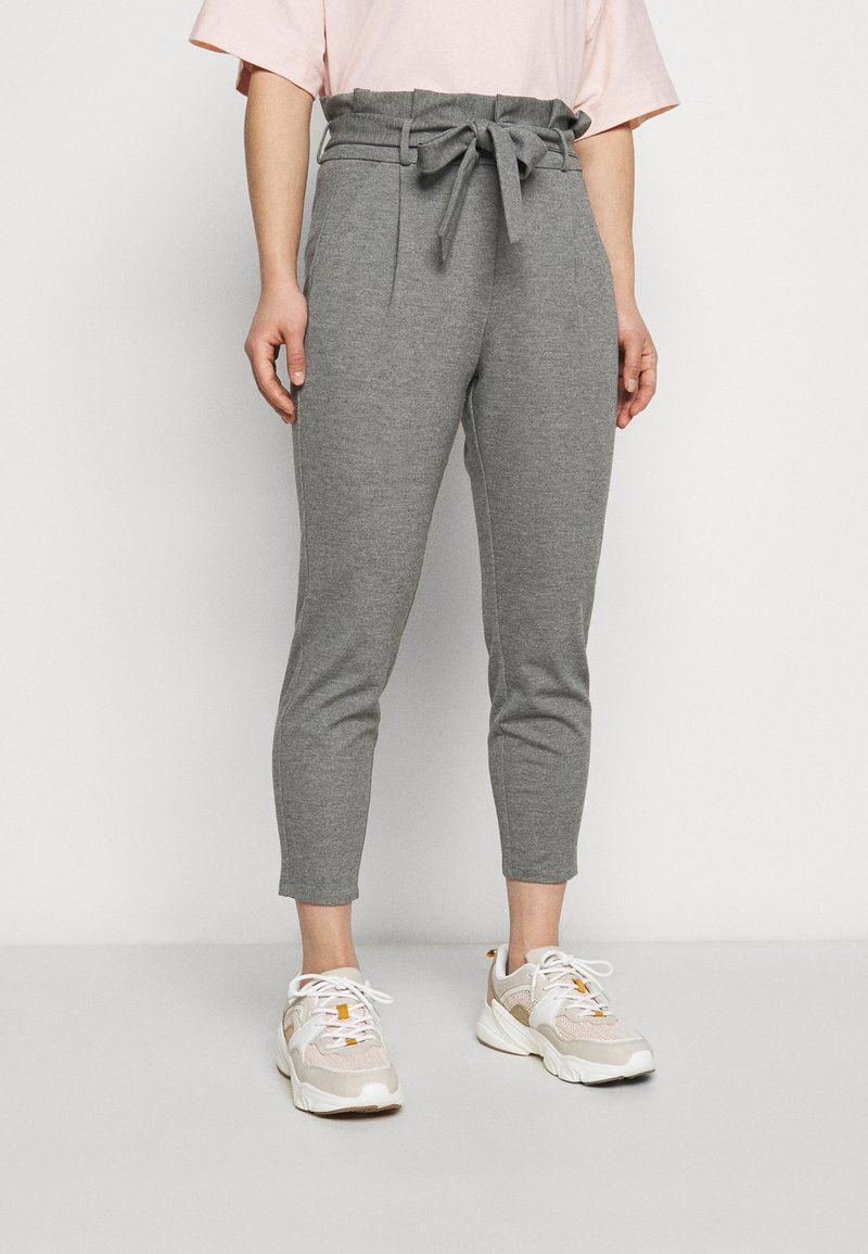 Žena v šedých kalhotách s vysokým pasem a zavázaným pásem, obutá v bílých teniskách, stojící na jednobarevném světlem pozadí.