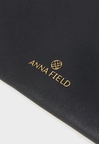 Juodas dirbtinės odos gaminys, featuring iškilmingą auksinį "ANNA FIELD" logotipą ir gėlių ikoną, demonstruojantis lygią tekstūrą ir stilingą dizainą.