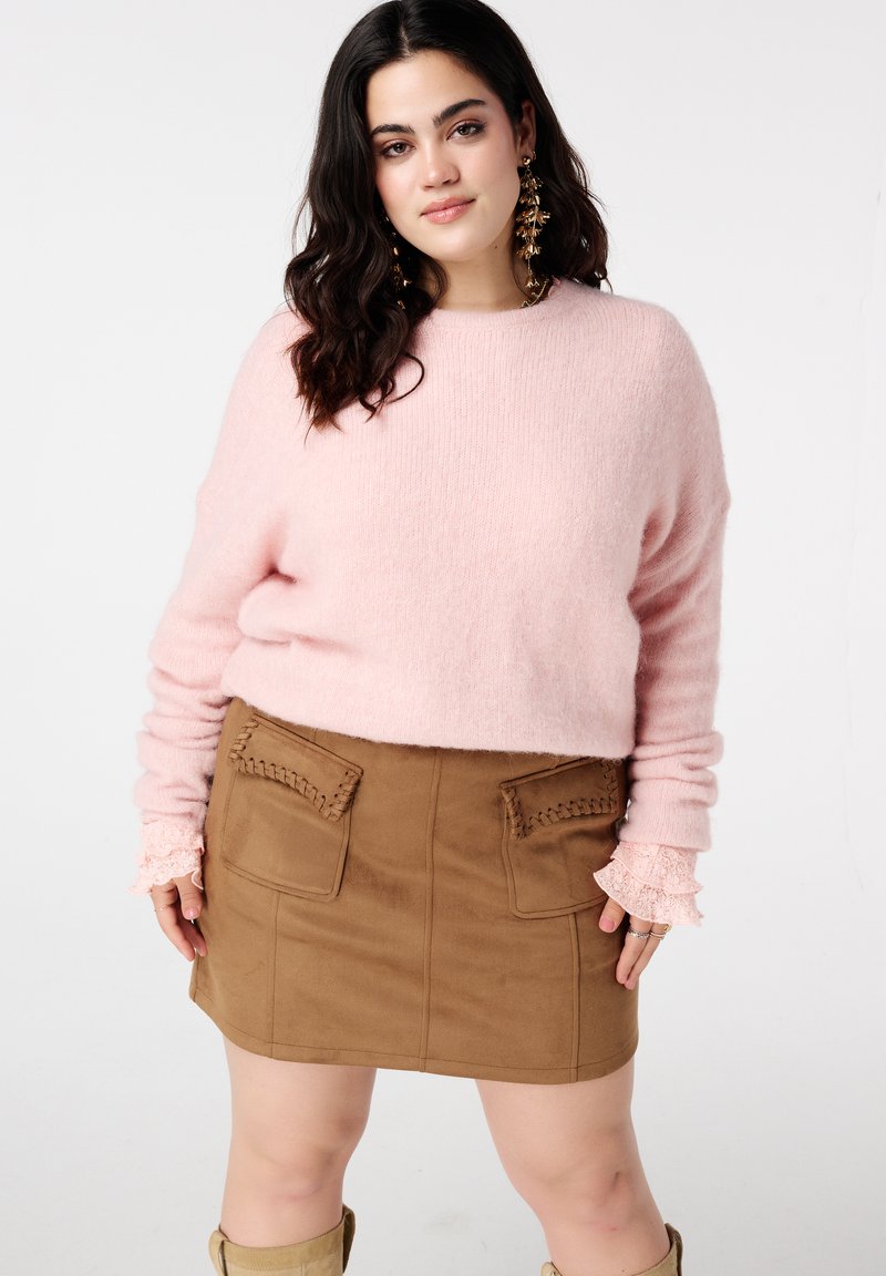 Pull en maille rose clair douce avec manches longues et poignets à volants, associé à une mini-jupe en suède marron avec poches cousues.